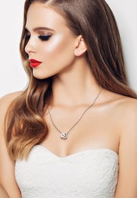 Sølv halskæde med en snoet, hjerteformet vedhæng båret på bar hud. Modellen har langt, bølget hår og en off-shoulder hvid top.