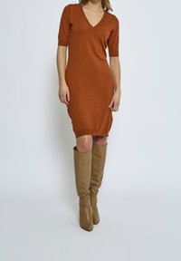 Robe en tricot couleur rouille avec un décolleté en V et des manches courtes, présentant une silhouette ajustée et un ourlet côtelé. Associée à des bottes en cuir beiges montantes.