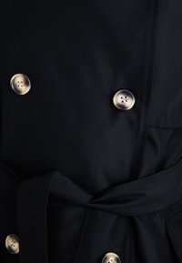 Fekete trench coat, amely dupla soros gombolású, aranyszínű gombokkal, szövet övvel és sima textúrával rendelkezik.