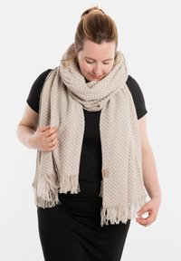 Lichtbeige geweven sjaal met een gestructureerd visgraatpatroon, franjes aan de randen en een klein merklabel, gewikkeld om de nek.