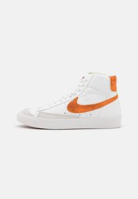 Höga sneakers med vit läderövre, orange mocka swoosh och grå gummitå. Har vita snören och varumärkeslogotyp på tungan.