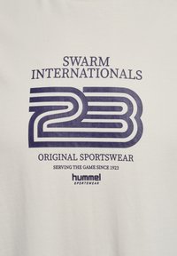 Witte t-shirt met een marineblauw graphic waarop de tekst "SWARM INTERNATIONALS 23," "ORIGINAL SPORTSWEAR," en "hummel SPORTSWEAR" logo's staat.
