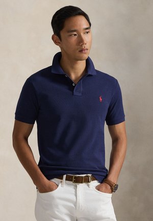 Mand iført en navyblå polo-shirt med en rød logo, hvide bukser, brun bælte og armbåndsur, stående med hænderne i lommerne mod en neutral baggrund.