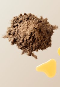 Brun pulverhög på ljus bakgrund, med två gula droppar i närheten. Texturerad yta med fina korn och släta former.