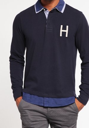 Mand iført en langærmet navy polo med en blå krave og hvid "H" emblem, kombineret med grå bukser og hænderne i lommerne.