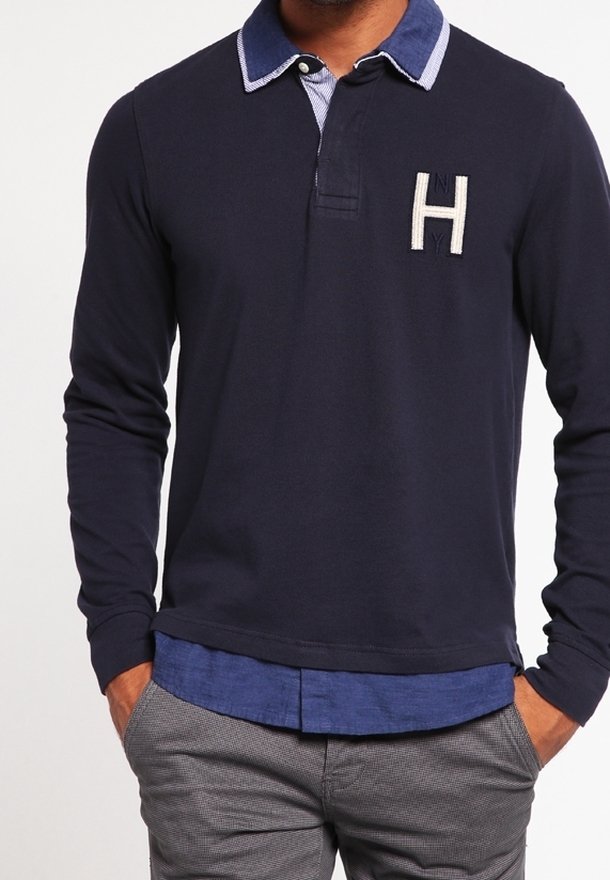 Homme portant un polo bleu marine à manches longues avec un col bleu et un emblème blanc "H", associé à un pantalon gris et les mains dans les poches.