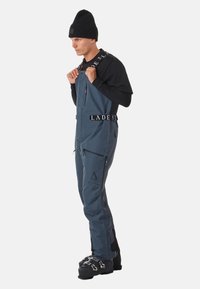 Marineblaue wasserdichte Schneehosen mit verstellbaren Trägern, Reißverschlusstaschen und schwarzen Akzenten. Getragen über einem langen schwarzen Shirt und einer Mütze.
