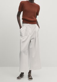 Maglione all'uncinetto color ruggine con maniche corte, abbinato a pantaloni bianchi a gamba larga. Le calzature presentano delicate fasce bianche.