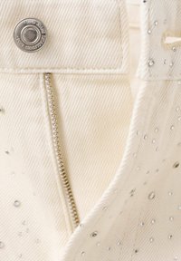 Gros plan sur un tissu en denim couleur crème avec une fermeture éclair argentée, un bouton en métal et de petites clous argentés dispersés sur le matériau.