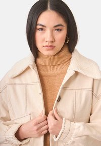 Chaqueta de mezclilla beige con cuello de borreguito, cuenta con bolsillos frontales y costuras visibles, superpuesta a un suéter de cuello alto marrón con relieves.