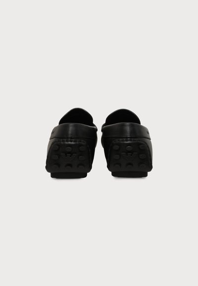 Ζευγάρι μαύρα δερμάτινα παπούτσια τύπου loafers χωρίς κορδόνια, εμφανιζόμενα από πίσω με ελαστικές σόλες και ένα μικρό ανάγλυφο λογότυπο σε κάθε τακούνι.