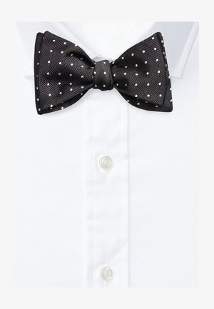 Melnā satīna tauriņš ar baltām pumpiņām, ar teksturētu virsmu, eleganti sasiets, uz baltas krekla apkakles fona.