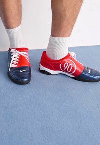 Nohy v bielych ponožkách a červených a tmavomodrých topánkach Nike s bielymi šnúrkami, s logom "90" na vonkajšej strane pravej topánky.