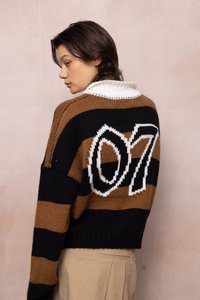 Noor inimene, kellel on lühikesed tumedad juuksed, on riietatud pruunikasmustrilisse triibulisse sviitresse, mille seljal on valge number "07", ja beežidesse pükstesse, olles veidi vasakule pöördunud.