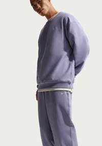 Sweat-shirt violet et pantalon assorti en tissu doux, avec un logo Nike blanc. Le modèle présente une coupe décontractée et des poignets côtelés.