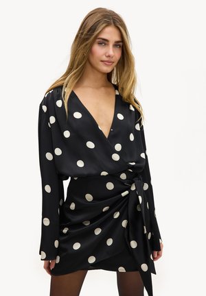 Femme aux longs cheveux blonds portant une robe portefeuille noire à grands pois blancs, posant devant un fond blanc uni.