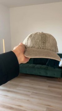 Șapcă bej cu material texturat, vizieră curbată și detalii cusute. Ținută în mână pe un fundal neutru, cu o canapea verde.