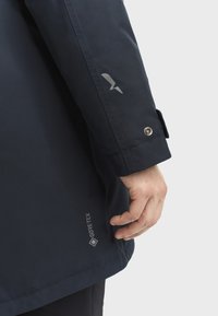 Mornarska vodootporna jakna s oznakom GORE-TEX, srebrnim logotipom na rukavu i zatvaračem na manžetama. Glatka tekstura, prilagođeni kroj.