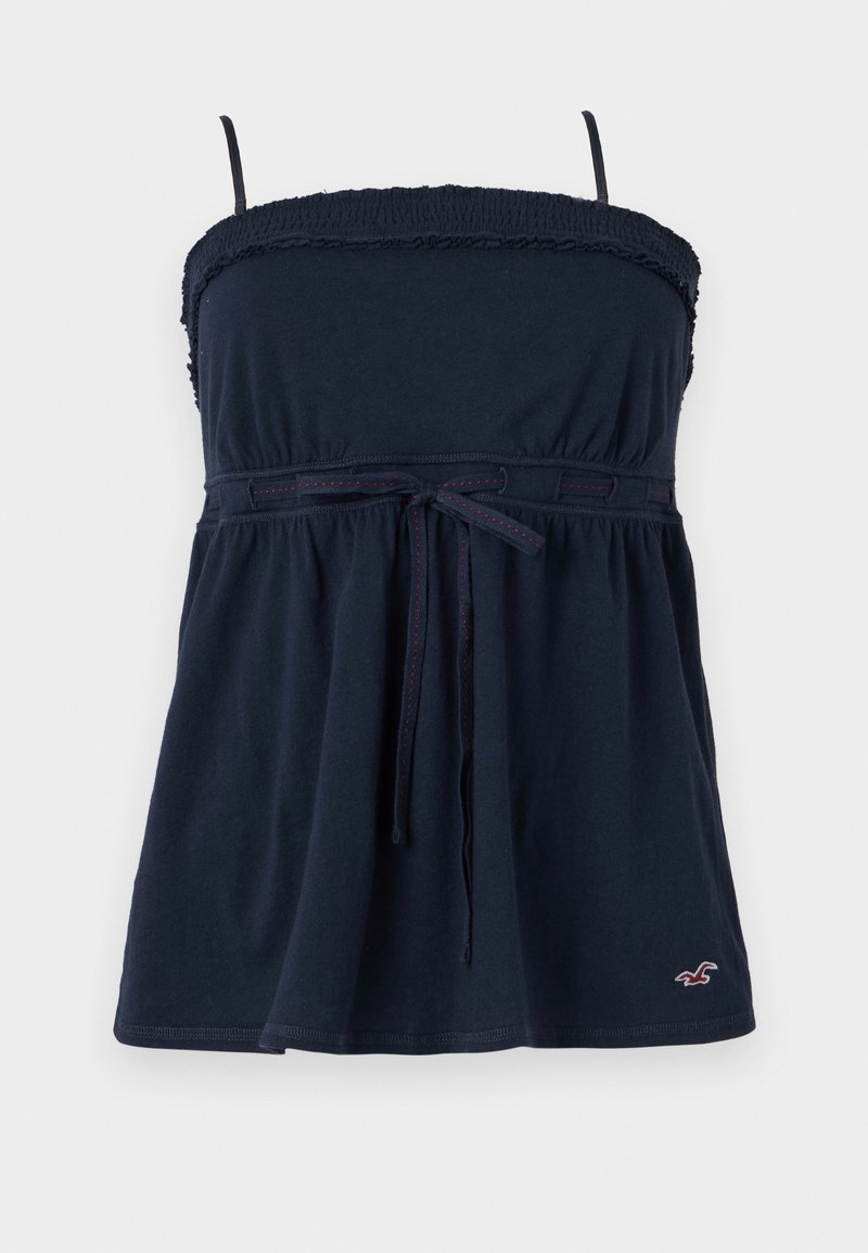 Top sin mangas de color azul marino con escote elástico fruncido, cintura con lazo y dobladillo acampanado. Presenta un sutil bordado rojo y un pequeño logo en la parte inferior.