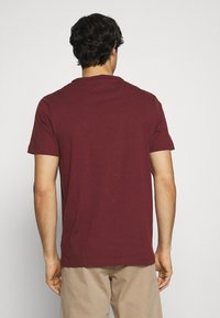 Uomo che indossa una semplice t-shirt corta bordeaux e pantaloni beige, in piedi con le spalle alla macchina fotografica su uno sfondo bianco.
