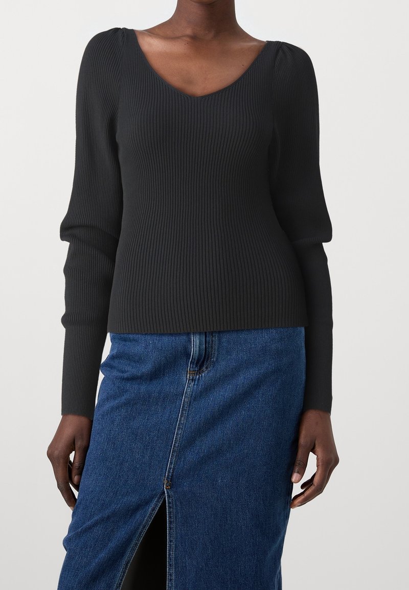 Femme portant un haut noir côtelé à manches longues avec col en V et une jupe en denim bleue avec fente devant, debout devant un fond uni.