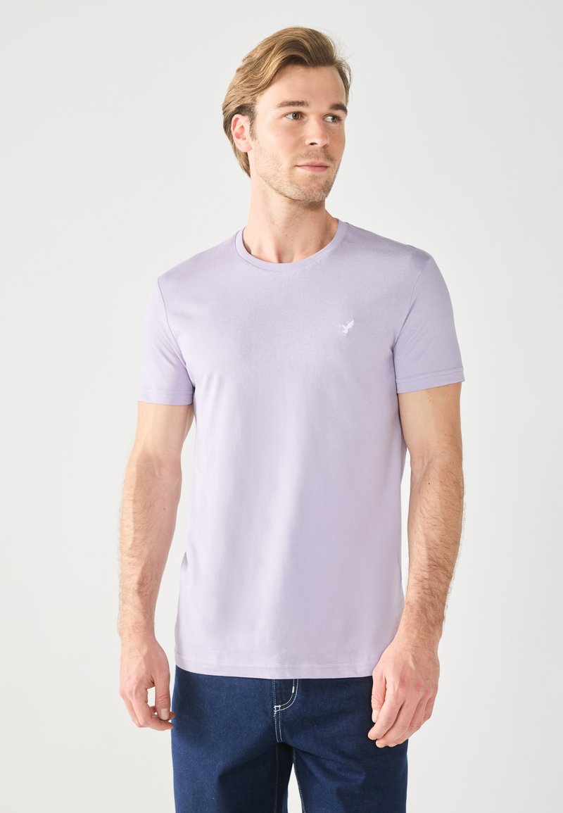 Tricou mov deschis cu guler rotund, fabricat din bumbac moale; are mâneci scurte și un logo mic, discret pe piept. Asortat cu blugi negri.