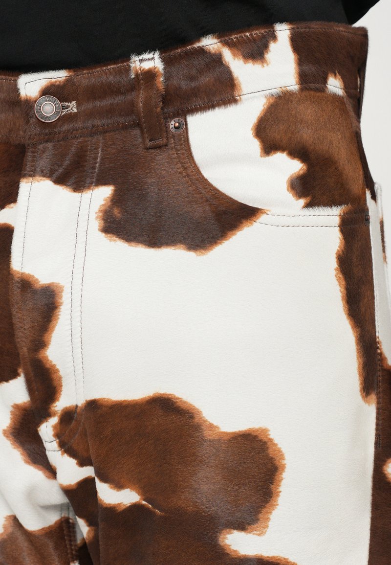 Jeans mit Kuhmuster in Braun und Weiß, aus weichem Stoff gefertigt. Enthalten sind Standardtaschen und ein Metallknopf am Bund.