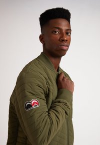 Giacca bomber trapuntata verde oliva con colletto e polsini a coste. La manica sinistra presenta una toppa multicolore con grafica di montagne.