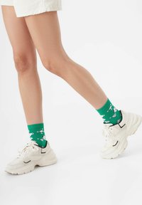 Grüne Socken mit einem verspielten Muster aus weißen cartoonhaften Tieren, kombiniert mit klobigen weißen Turnschuhen mit schwarzen Akzenten und strukturierten Details.