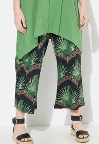 Top verde combinado con pantalones negros con estampado de hojas tropicales; sandalias negras con tira en el tobillo y suela de esparto.
