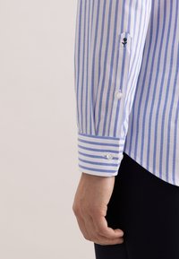 Camicia bianca con righe blu, caratterizzata da polsino con bottoni e piccolo dettaglio con logo ancora sulla manica. Tessuto dalla texture liscia.