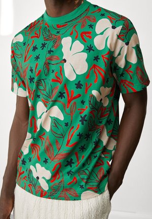 Kurzärmeliges T-Shirt aus grünem Baumwollstoff mit einem floralen Muster in Weiß, Rot und Marineblau, das geschwungene Formen und Blattmotive zeigt.