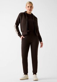 Schwarze Zip-up-Jacke mit weißen Seitenstreifen, kombiniert mit passendem schwarzem Crop-Top und zulaufender Hose. Weiße Sneakers vervollständigen das Outfit.