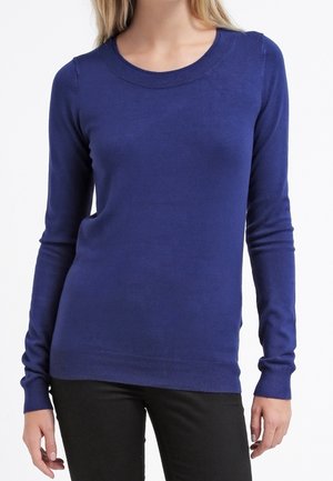 Femme portant un pull bleu marine ajusté à manches longues et col rond, associé à un pantalon noir sur fond blanc.