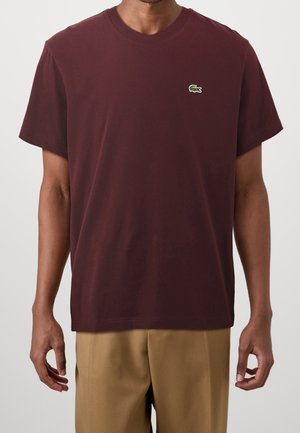 Mann trägt ein burgunderrotes T-Shirt mit kurzen Ärmeln und einem kleinen grünen Krokodil-Logo auf der linken Brust sowie beige Hosen.