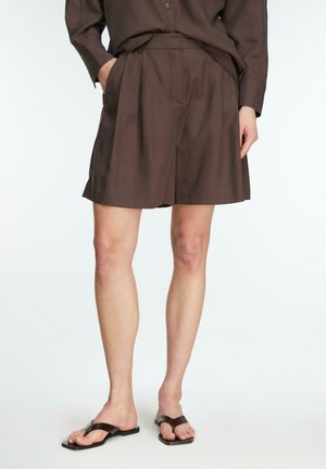 Brązowe plisowane szorty z wysokim stanem i bocznymi kieszeniami, noszone z dopasowaną koszulą z długim rękawem i ciemnobrązowymi sandałami z odkrytymi palcami.