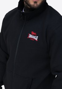 Felpa nera con zip realizzata in un materiale morbido, dotata di un colletto alto, tasche frontali e un logo di leone ricamato in rosso sul petto.