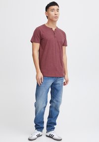 Camisa henley de manga corta color marrón con una tapeta de tres botones, combinada con unos jeans de mezclilla rectos en azul claro y zapatillas deportivas en blanco y negro.