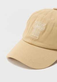 Μπεζ καπέλο μπέιζμπολ με κεντητό λευκό σχέδιο κλασικής κολώνας στην μπροστινή όψη, καμπύλη γείσο και ραμμένες τρύπες αερισμού.