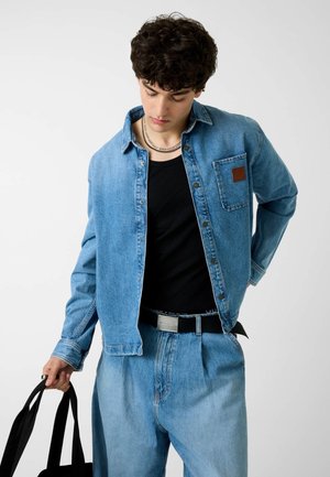 Jeune homme portant une veste en jean et un jean, un T-shirt noir, tenant un sac fourre-tout noir, regardant vers le bas sur un fond uni.