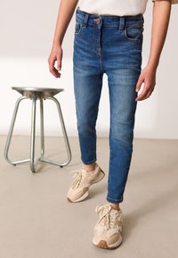 Visoko pasovane, slim fit modre kavbojke z zbledelim videzom, v kombinaciji s bež športnimi copati, ki imajo teksturirane detajle in obloženo podplat.