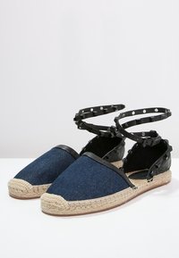 Blå denim espadriller med svarta läderdetaljer, jutesulor och ankelremmar med metallisk nitar. Öppen bak för enkel påtagning.