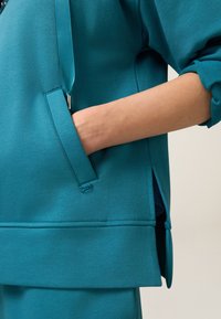 Sudadera en color azul verdoso con un bolsillo lateral, un detalle de cremallera y un corte relajado con mangas con puños. Tejido suave con una textura blanda.