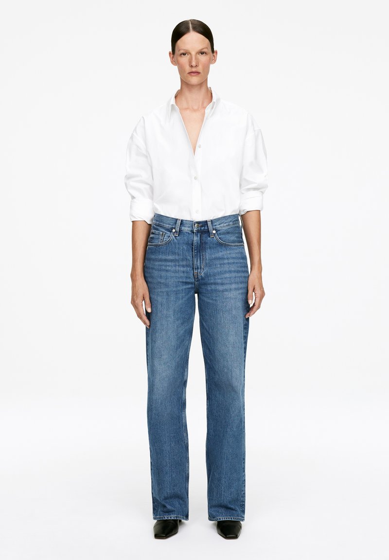 Chemise blanche à boutons, avec des manches retroussées, associée à un jean en denim flared taille haute dans un délavage bleu moyen. La tenue est complétée par des chaussures noires.