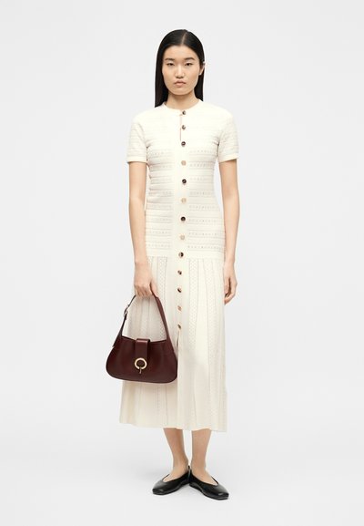 Γυναίκα που φοράει μπεζ μεσαίο φόρεμα με κουμπιά στο μπροστινό μέρος, κρατώντας μια σκούρα καφέ τσάντα, συνδυασμένο με μαύρα μπαλαρίνες, σε λευκό φόντο.