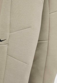 Beige Sporthosen aus strukturiertem Material mit schrägen Seitentaschen und einem kleinen schwarzen Logo auf der unteren linken Seite.