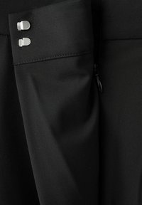 Ärmel eines schwarzen Kleidungsstücks mit zwei silbernen, rechteckigen Manschettenknöpfen und einem teilweise sichtbaren schwarzen Reißverschluss auf strukturiertem Stoff.