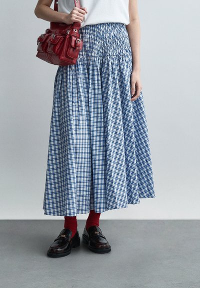 Vue de la taille vers le bas d'une personne portant une longue jupe à carreaux bleus et blancs, des chaussettes rouges, des mocassins noirs, tenant un sac à main rouge avec des détails de boucle.