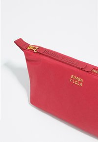 Neceser de nylon rojo con herrajes dorados, que cuenta con cierre de cremallera y logotipo grabado. Textura suave con un acabado mate, forma cónica.