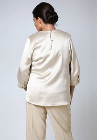 Woman with dark hair in a bun wearing a cream satin blouse with back button detail and matching beige trousers, standing against a plain background.
